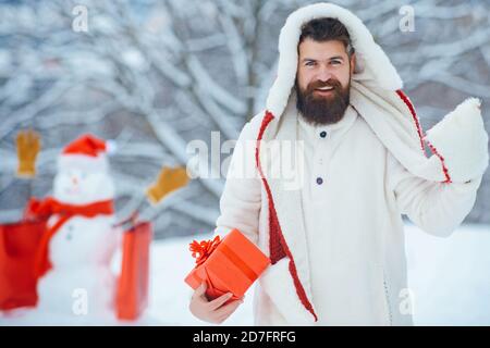 Lustige Santa Mann posiert mit roten Geschenkbox auf Winterwetter. Fröhlicher Vater halten Geschenk und Spaß mit Schneemann im Winter Park. Stockfoto