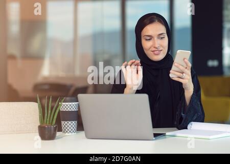 Glückliche muslimische Geschäftsfrau in Hijab am Büroarbeitsplatz. Lächelnde arabische Frau, die am Laptop arbeitet und mit dem Smartphone spricht Stockfoto