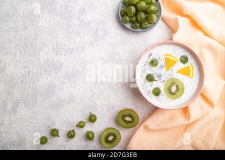 Joghurt mit Kiwi, Stachelbeere, Chia in Keramikschale auf grauem Betongrund und orangefarbenem Leinentextilien. Draufsicht, flaches Lay, Kopierraum. Stockfoto