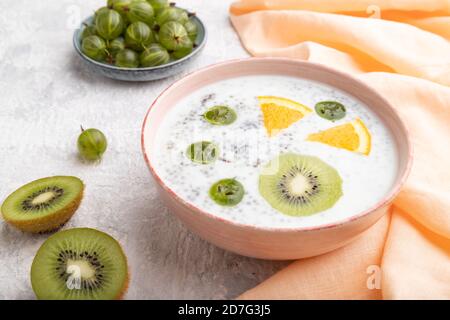 Joghurt mit Kiwi, Stachelbeere, Chia in Keramikschale auf grauem Betongrund und orangefarbenem Leinentextilien. Seitenansicht, Nahaufnahme. Stockfoto