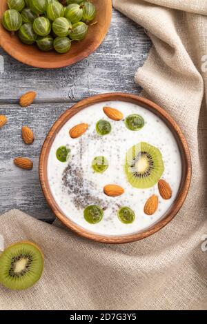 Joghurt mit Kiwi, Stachelbeere, Chia und Mandeln in Holzschüssel auf grauem Holzhintergrund und Leinentextilien. Draufsicht, flaches Lay, Nahaufnahme. Stockfoto