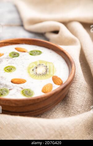 Joghurt mit Kiwi, Stachelbeere, Chia und Mandeln in Holzschüssel auf grauem Holzhintergrund und Leinentextilien. Seitenansicht, Nahaufnahme, selektiver Fokus. Stockfoto