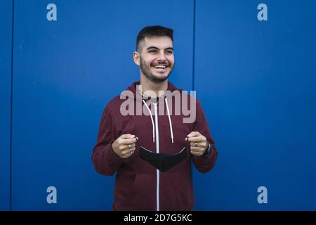 Fröhlich lächelnd jungen Mann trägt eine rote Jacke und Putting Auf einer Gesichtsmask - COVID-19 Stockfoto