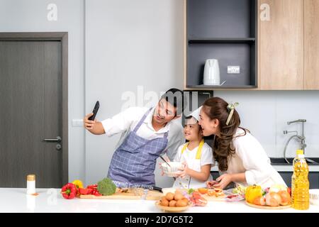 Junge asiatische Familie bereiten Salat in der Küche und Vater nehmen ein Foto Selfie per Telefon. Aufgeregt lächeln und Fällen glücklich. Eltern lehren Tochter Stockfoto