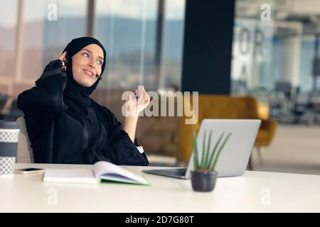 Glückliche muslimische Geschäftsfrau in Hijab am Büroarbeitsplatz. Lächelnde arabische Frau, die am Laptop arbeitet und mit dem Smartphone spricht Stockfoto