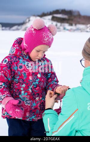 Ein Mädchen in einer grünen Jacke legt einem kleinen Mädchen einen Fäustling auf die Hand, während es im Winter im Park spazieren geht. Pflege der Hände im Winter. Schutz von Stockfoto