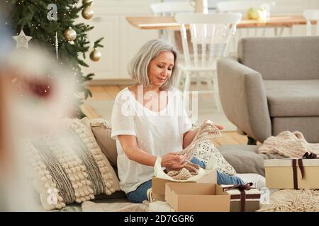 Reife Frau hält warmen Schal in den Händen beim Öffnen Geschenke während Heiligabend zu Hause Stockfoto
