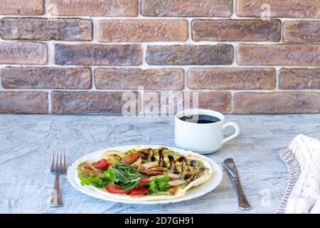 Italienisches Frühstück mit Kaffee. Piadina mit gegrilltem Hähnchen, Tomaten, Salat und Rucola. Köstliches Frühstück auf dem Tisch mit Serviette und Backsteinwand Hintergrund serviert. Weichfokus Stockfoto