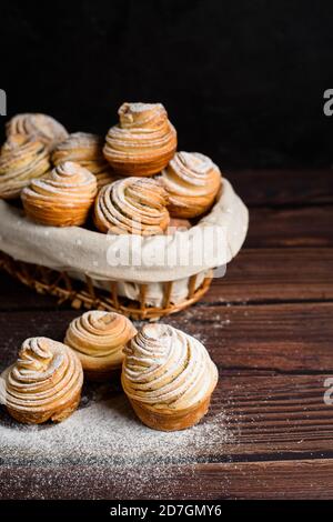 Leckere moderne Cruffins werden aus Puffhörnchen in Form eines Muffins hergestellt. Cruffins mit einem Holzkorb auf dunklem Holzhintergrund, bestreut Stockfoto