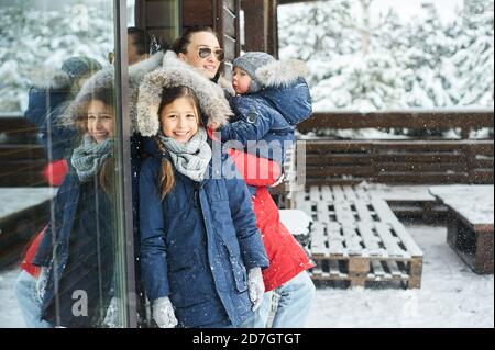 Ein Portret der schönen jungen kaukasischen Mutter mit ihren Kindern Im Winter in der Nähe des Hauses mit einem schneebedeckten weihnachten Bäume Stockfoto