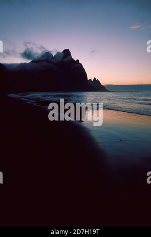 Eine minimalistische dunkle Berg- und Strandlandschaft mit niedriger Stimmungslage bewölkt und Himmel - Vestrahorn Berg und der schwarze Sand Strand in Stokksnes Island Stockfoto