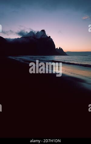 Eine minimalistische dunkle Berg- und Strandlandschaft mit niedriger Stimmungslage bewölkt und Himmel - Vestrahorn Berg und der schwarze Sand Strand in Stokksnes Island Stockfoto