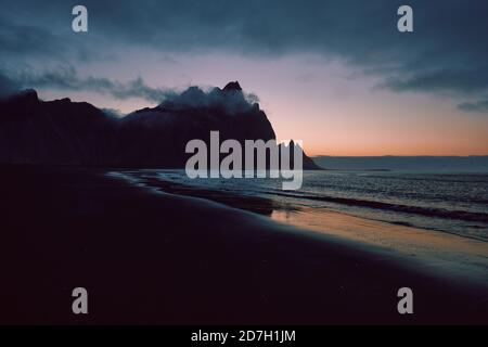 Eine minimalistische dunkle Berg- und Strandlandschaft mit niedriger Stimmungslage bewölkt und Himmel - Vestrahorn Berg und der schwarze Sand Strand in Stokksnes Island Stockfoto