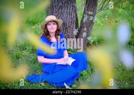 Eine Frau in einem Strohhut sitzt mit einem Buch Unter einem Baum im Frühlingswald Stockfoto