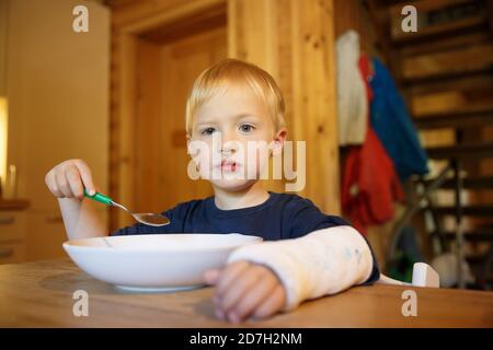 Kleiner Junge mit gebrochenem Handgelenk beim Essen am Tisch. Junge mit einem Gips auf der Hand. Stockfoto