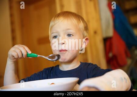 Kleiner Junge mit gebrochenem Handgelenk beim Essen am Tisch. Junge mit einem Gips auf der Hand. Stockfoto