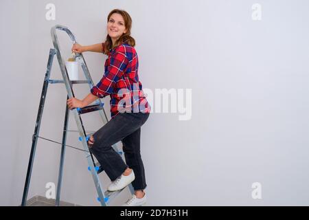 Eine Malerin steht auf einer Trittleiter mit Pinsel und einer Dose Farbe, Raum kopieren. Malen der Wände in einer neuen Wohnung Stockfoto