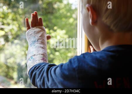 Kleiner Junge mit Gips am Arm, der durch das Fenster schaut und die Natur beobachtet. Stockfoto