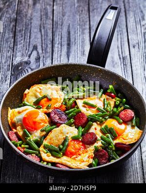 Spanisches Frühstück mit grünen Bohnen gebackenen Eiern und würzigen gebratenen Chorizo-Wurst auf einer Pfanne auf einem Holzhintergrund, Draufsicht, Nahaufnahme Stockfoto