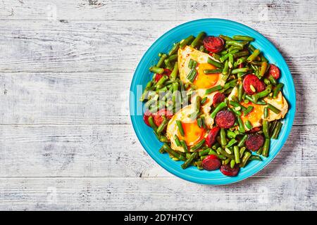 Spanisches Frühstück mit grünen Bohnen gebraten sonnigen Seite up Eier und würzig gebratenen Chorizo Wurst auf einem blauen Teller mit Besteck auf einem Holztisch, Draufsicht, cl Stockfoto