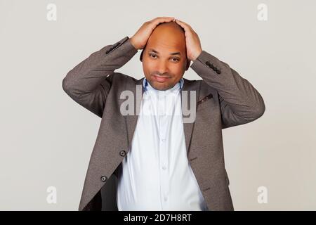Porträt eines jungen afroamerikanischen Mannes Geschäftsmann, hält seine Hände auf dem Kopf, müde Gesichtsausdruck wegen Kopfschmerzen nach einem harten Tag bei der Arbeit. Stehen auf einem grauen Hintergrund Stockfoto