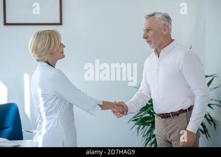Grauhaarige männliche Patientin schüttelt sich die Hände mit blonden weiblichen Arzt Stockfoto