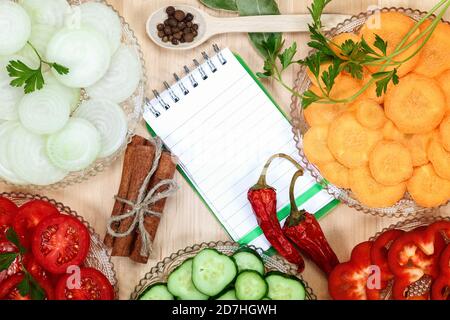 Frisches, gehacktes Gemüse, Gewürze und ein Notizbuch mit Rezepten auf einem Holzschneidetisch. Stockfoto