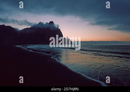 Eine minimalistische dunkle Berg- und Strandlandschaft mit niedriger Stimmungslage bewölkt und Himmel - Vestrahorn Berg und der schwarze Sand Strand in Stokksnes Island Stockfoto