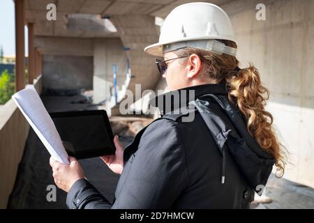 Rückansicht einer jungen Architektin, die einen Schutzhelm trägt, Papierbodenpläne hält und ein digitales Gerät auf einer Baustelle überprüft Stockfoto