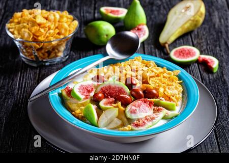 Gesundes Frühstück: Blaue Schale mit naturbelassenem Joghurt mit knusprigen Maisflocken, Feigen, Birne auf schwarzem Holzgrund serviert, Draufsicht, clos Stockfoto