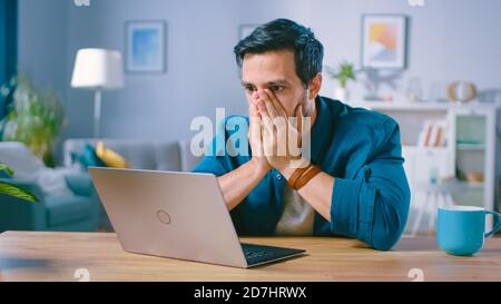Aufnahme eines verärgerten jungen Mannes bedeckt sein Gesicht mit Palms in Frustration, während er schlechte Nachrichten vom Lesen schlechter Nachrichten auf einem Laptop erhält. Verärgert Mann zu Hause. Stockfoto
