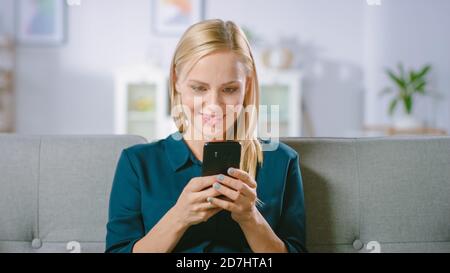 Schöne Blonde Frau nutzt Handy, während auf einem Sofa zu Hause sitzen. Smiling Happy Woman nutzt Smartphone für das Surfen im Internet, Social Stockfoto