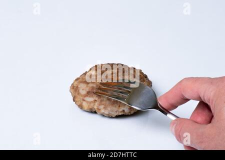 Hand hält eine Gabel über gekochten Fleisch Patty auf einem weißen Teller isoliert auf weißem Hintergrund, Nahaufnahme, Kopierraum Stockfoto
