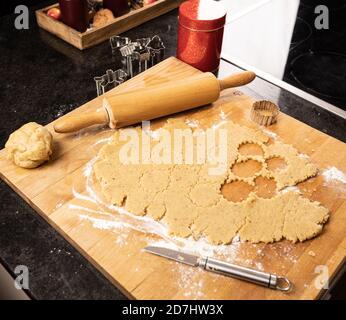 Plätzchenteig (Mürbeteig) auf Mehl auf einem hölzernen Küchentafel auf schwarzer Theke ausgerollt. Nudelholz, Ausstechformen und Blechdose sichtbar. Runde Kochstellen Stockfoto