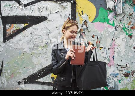 Glückliche Frau, die ein Buch auf der Straße in der Nähe von Graffiti liest An der Wand Stockfoto