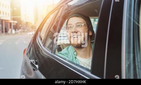 Stilvoller junger Mann fährt auf einem Beifahrer-Rücksitz eines Autos, schaut aus dem Fenster in Wonder. Großer sonniger Blick auf die Stadt spiegelt sich im Fenster. Schuss Gemacht Stockfoto