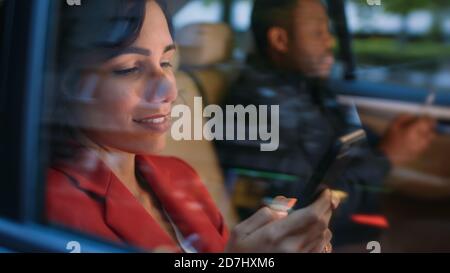 Junge Frau und Mann reiten am Abend auf dem Rücksitz eines Autos. Beide nutzen Smartphones, Browsen im Internet und machen Business Work. Kameraaufnahme Stockfoto