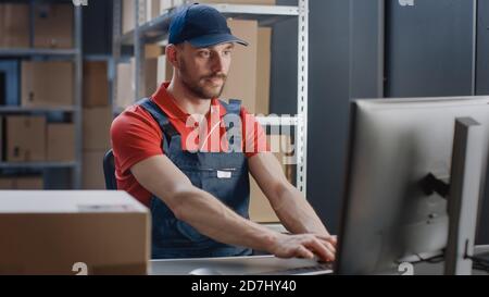Porträt eines uniformierten Arbeiters, der einen Computer benutzt, während er an seinem Schreibtisch im Lager sitzt. Stockfoto