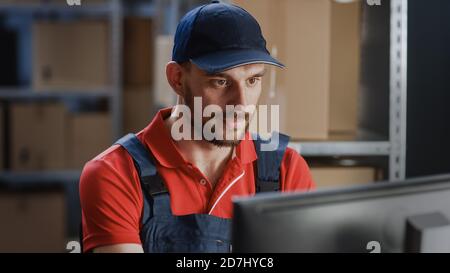 Porträt eines uniformierten Arbeiters, der einen Computer benutzt, während er an seinem Schreibtisch im Lager sitzt. Stockfoto