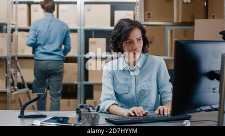 Weibliche Inventory Manager sitzt an ihrem Schreibtisch und mit Personal Computer, Arbeiter legt Paket auf dem designierten Regal. In den Zeilen Hintergrund von Stockfoto