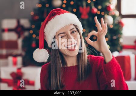 Schöne Mädchen heben Arm demonstrieren okey schließen Auge tragen santa Kopfbedeckung roter Pullover in dekorierten Weihnachten Wohnzimmer drinnen Stockfoto