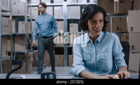 Weibliche Inventory Manager sitzt an ihrem Schreibtisch und mit Personal Computer, Arbeiter legt Pakete auf dem designierten Regal. In den Zeilen Hintergrund von Stockfoto