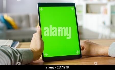 Nahaufnahme des Menschen mit Handgesten auf dem grünen Mock-up-Bildschirm Digital Tablet Computer im Portrait-Modus, während er an seinem Schreibtisch sitzt. Im Hintergrund gemütlich Stockfoto