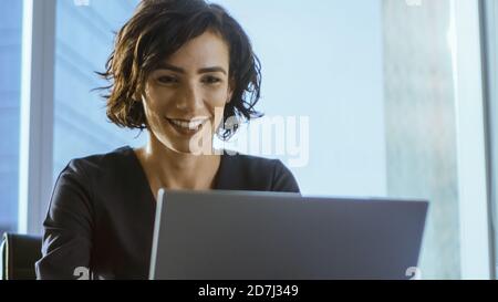 Selbstbewusste weibliche Führungskraft arbeitet auf einem Laptop sitzen an ihrem Schreibtisch in modernen Büro mit Blick auf die große Stadt. Lächelnd Erfolgreiche Busiesswoman Verwendet Laptop. Stockfoto