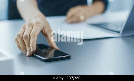 Aufnahme der Geschäftsfrau, die an ihrem Office Desk arbeitet und nach ihrem Smartphone greift. Frau nimmt Handy von ihrem Schreibtisch. Konzentrieren Sie sich auf ein Telefon. Stockfoto