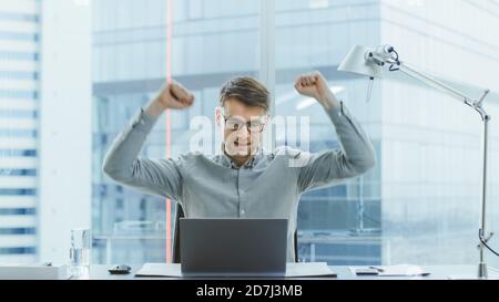 Handsome Young Businessman sitzt an seinem Schreibtisch mit Laptop im Büro hebt die Hände und feiert beeindruckende Finanzergebnisse. Im Hintergrund Stockfoto