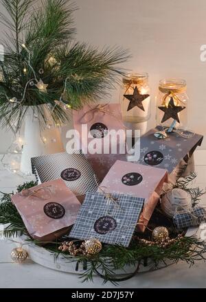 Adventskalender mit Paket auf einem weißen Holztablett, im Hintergrund ein Krug mit Kiefernästen bei warmem Kerzenlicht Stockfoto