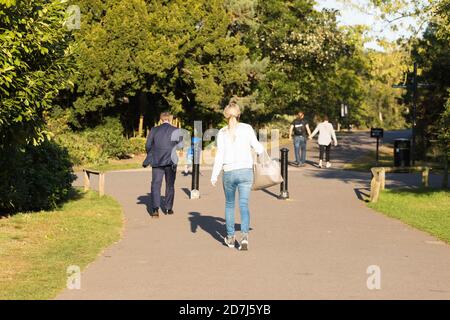 Verschiedene Passanten gehen an einem sonnigen Tag entlang des Asphaltpfades der Straße unter grünen Bäumen. Englischer Park. City People Style, Parkbereich, Frühherbst. Stockfoto