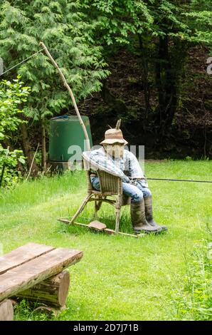 Vogelscheuche, aus Heu, Kleidung und Hut, lustige menschliche Figur, sitzt in einem Stuhl mit einer Angelrute in der Nähe eines Teiches in Deutschland, Westeuropa Stockfoto