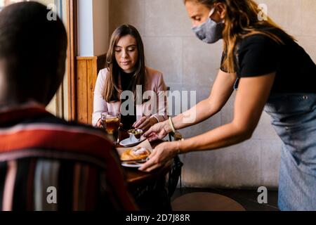 Kellnerin in schützende Gesichtsmaske serviert Essen zu jungen Paar Im Café während der COVID-19 Stockfoto